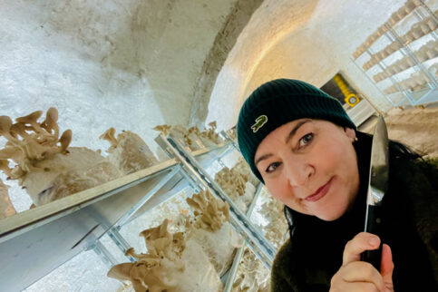 Martina Jansky steht mit einer Mütze und im dunklen Pullover, im Vordergrund des Bildes. Hinter ihr sieht man den Gewölbekeller in dem die Koenigspilze kultiviert werden. Rechts und links hinter ihr sieht man Metallregale mit Pilz-Substraten. auf einigen wachsen bereits Pilze die zur Ernte bereit sind. Martina Jansky hat ein Messer in der Hand, mit dem sie die Pilze nun ernten wird.