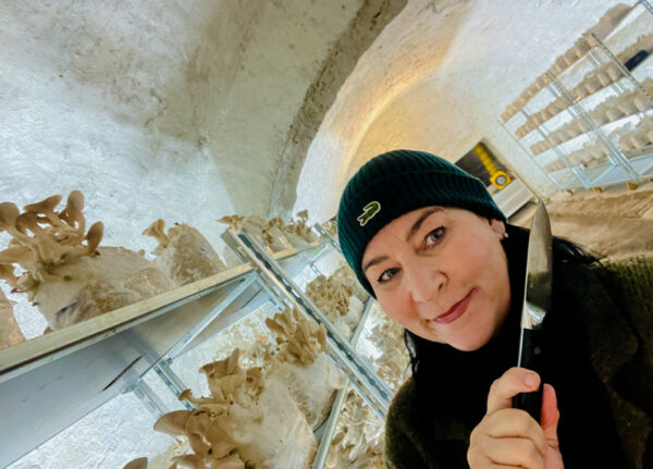 Martina Jansky steht mit einer Mütze und im dunklen Pullover, im Vordergrund des Bildes. Hinter ihr sieht man den Gewölbekeller in dem die Koenigspilze kultiviert werden. Rechts und links hinter ihr sieht man Metallregale mit Pilz-Substraten. auf einigen wachsen bereits Pilze die zur Ernte bereit sind. Martina Jansky hat ein Messer in der Hand, mit dem sie die Pilze nun ernten wird.